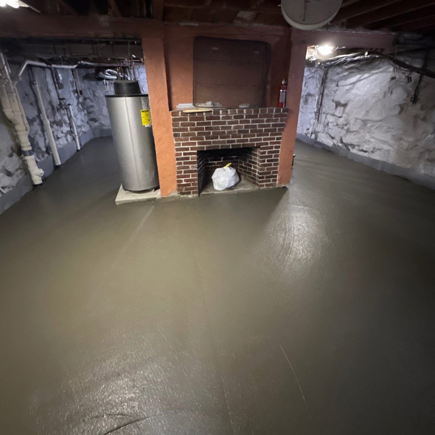 Basement with a fresh concrete floor. Brick fireplace, water heater, and stone walls.