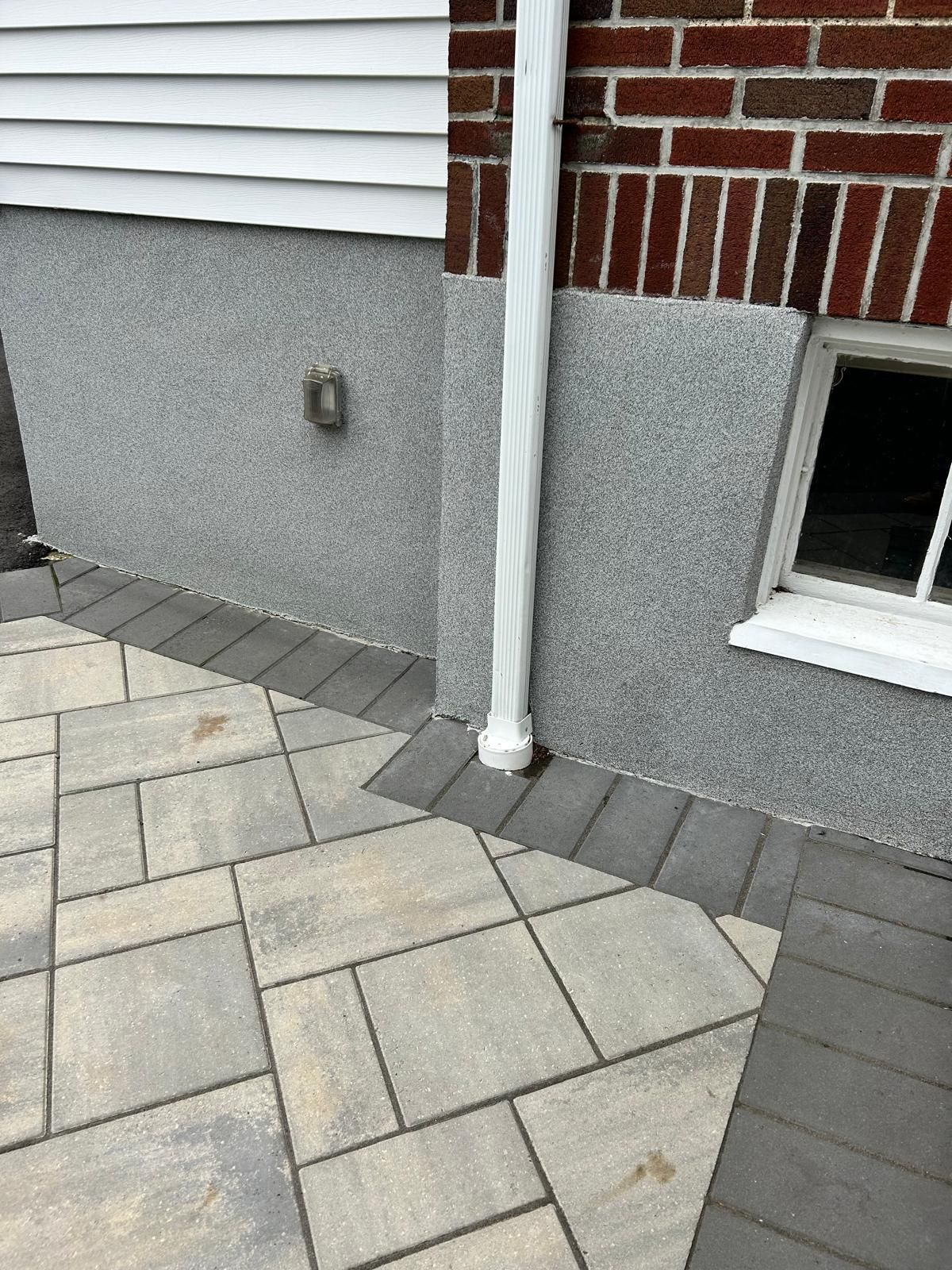 Exterior view of a house with gray stone veneer, white siding, and a paved patio. Brick wall with a window is to the right.