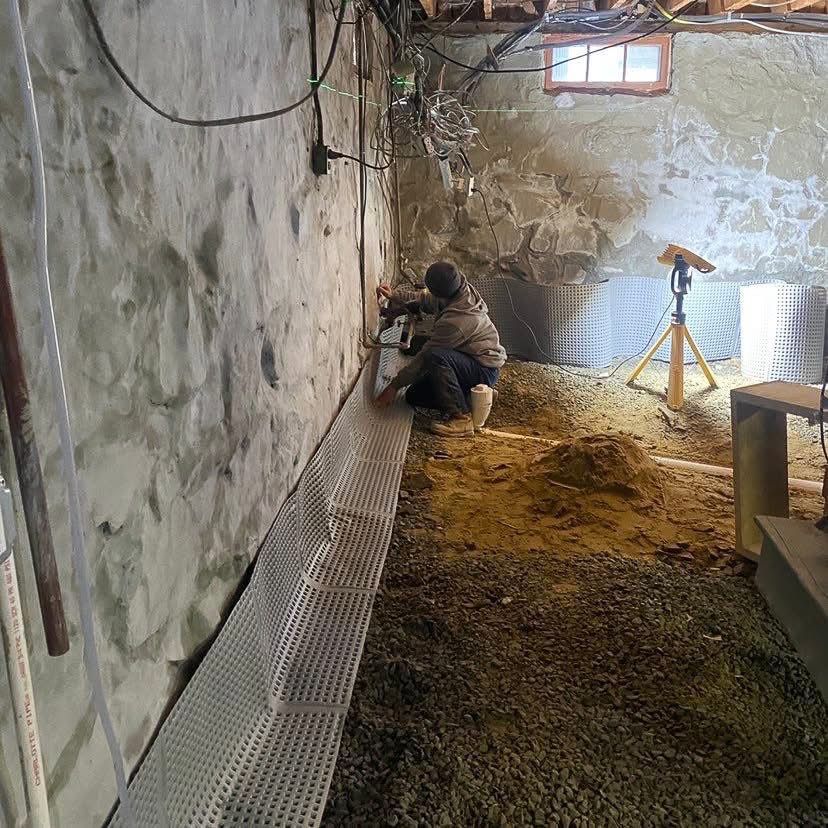 Person installing drainage against a stone basement wall. Gravel floor with a window and spotlight present.