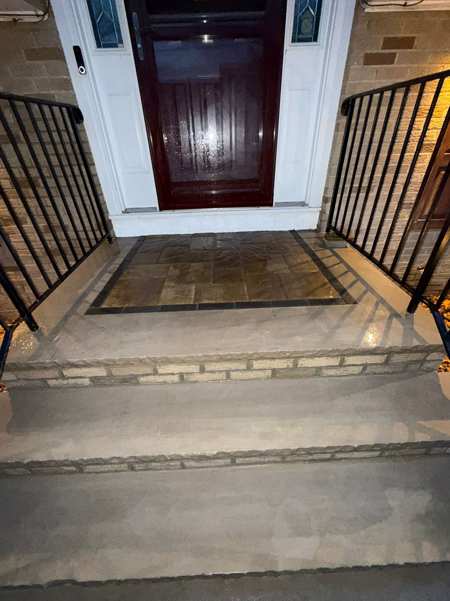 Exterior view of a house entrance with steps, railing, and door. Features brick accents, dark wood door, and patterned stone mat.