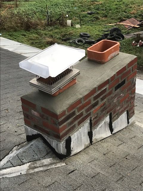 Brick chimney on a gray shingled roof. It has a chimney cap, terracotta pot, and concrete base.