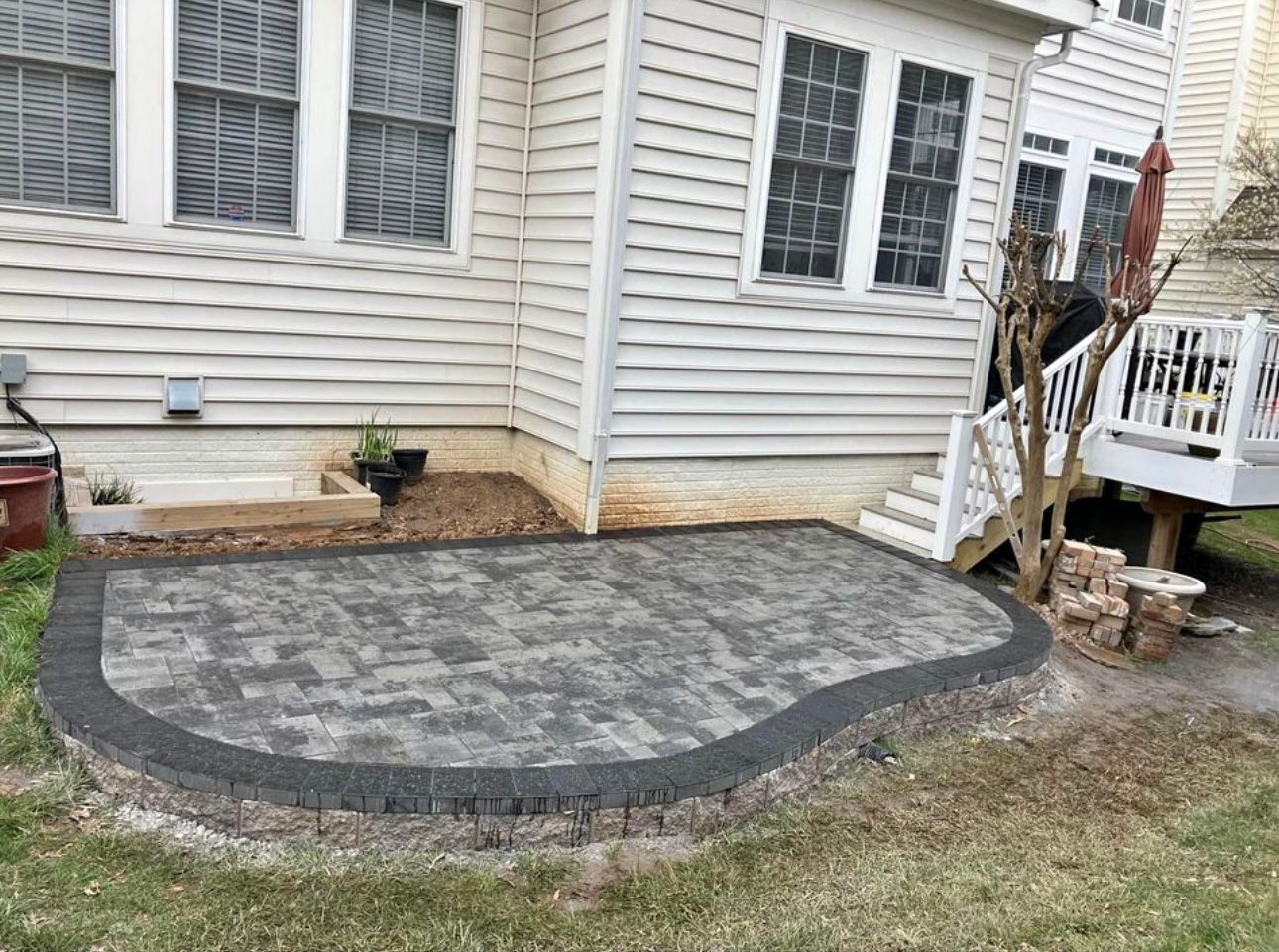 Backyard patio made of gray pavers, bordered by a dark gray stone wall, next to a house.