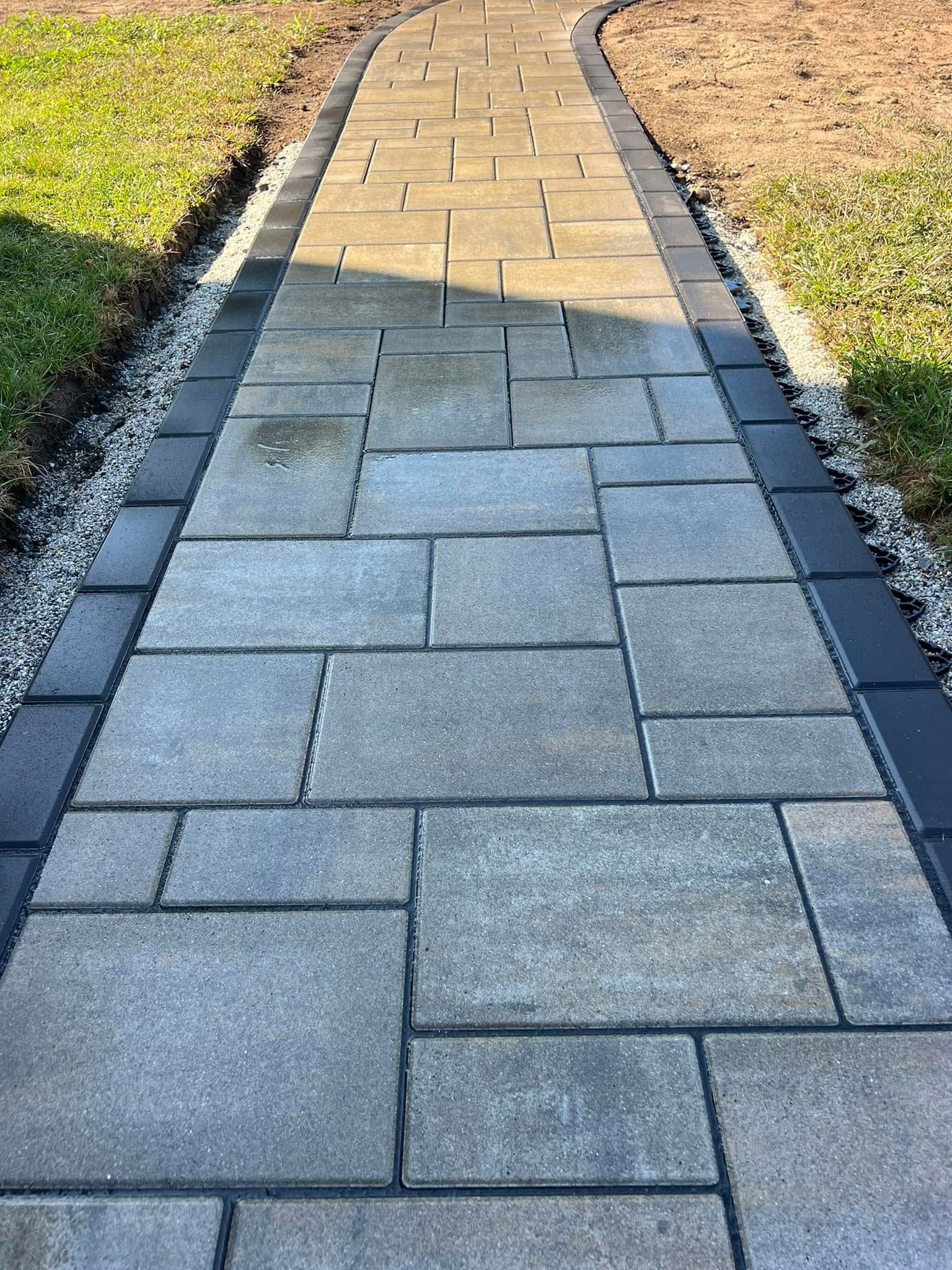 Stone pathway with a dark border, flanked by green grass and dirt.