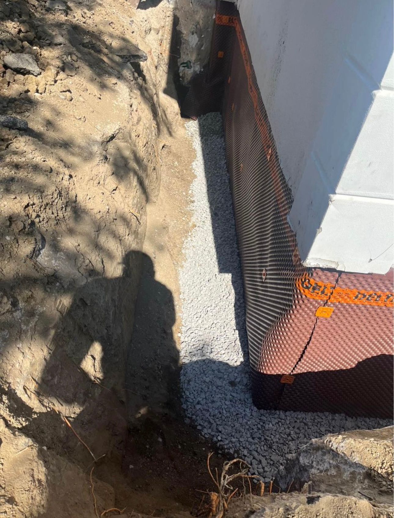 Trench filled with gravel next to a foundation wall covered with orange-accented black drainage mesh. Shadow of person.