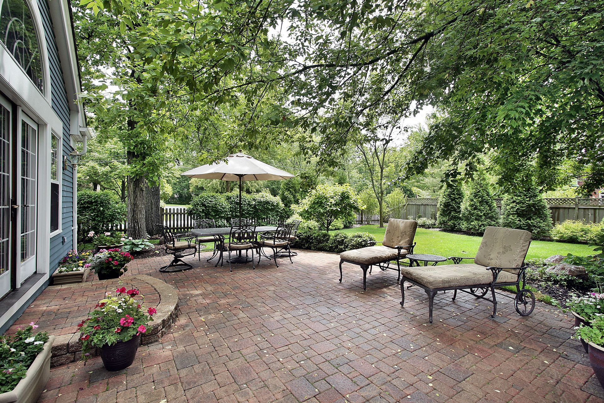 A brick patio with outdoor dining furniture, lounge chairs, and an umbrella, nestled in a lush, green, wooded backyard.