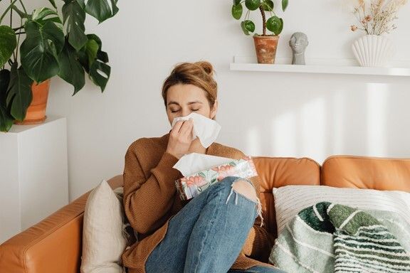 A woman is sitting on a couch blowing her nose into a napkin.