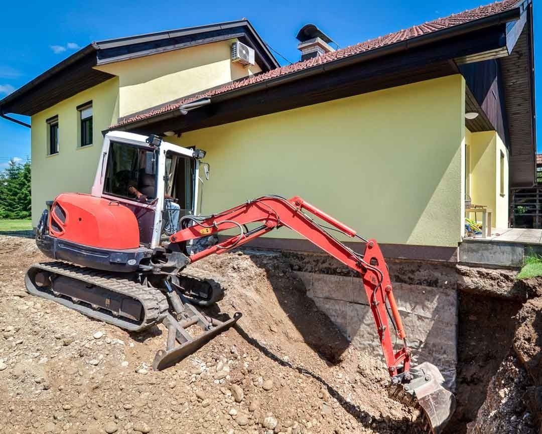 Red excavator digging next to a yellow house, outdoors.