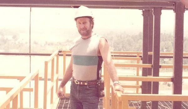 Man in hard hat, sleeveless shirt, and jeans on a construction site, holding onto a railing, looking towards the camera.
