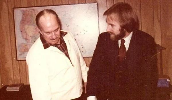 Two men in a room. One in a suit, the other in a jacket, looking at something. Map on the wall behind them.