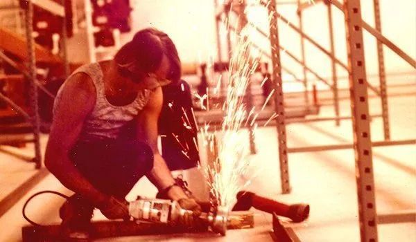 Person kneeling, grinding metal, sparks flying. In an industrial warehouse setting.
