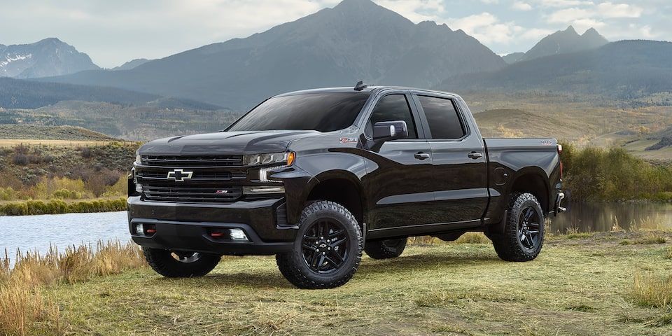 Black Chevrolet Silverado truck on a grassy field near water with mountains in the background.