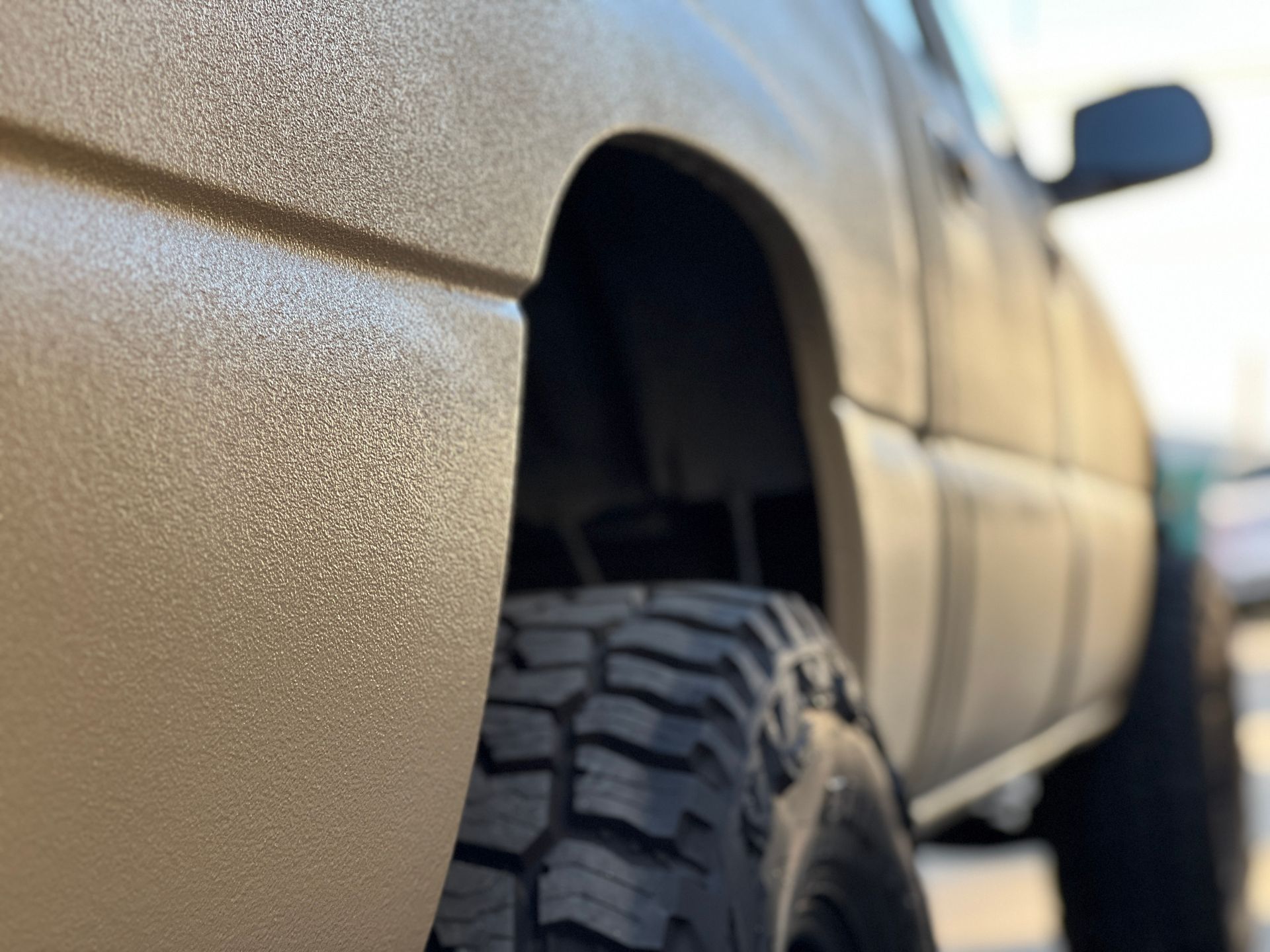 Tan pickup truck with textured paint, large black tire, and blurred background.