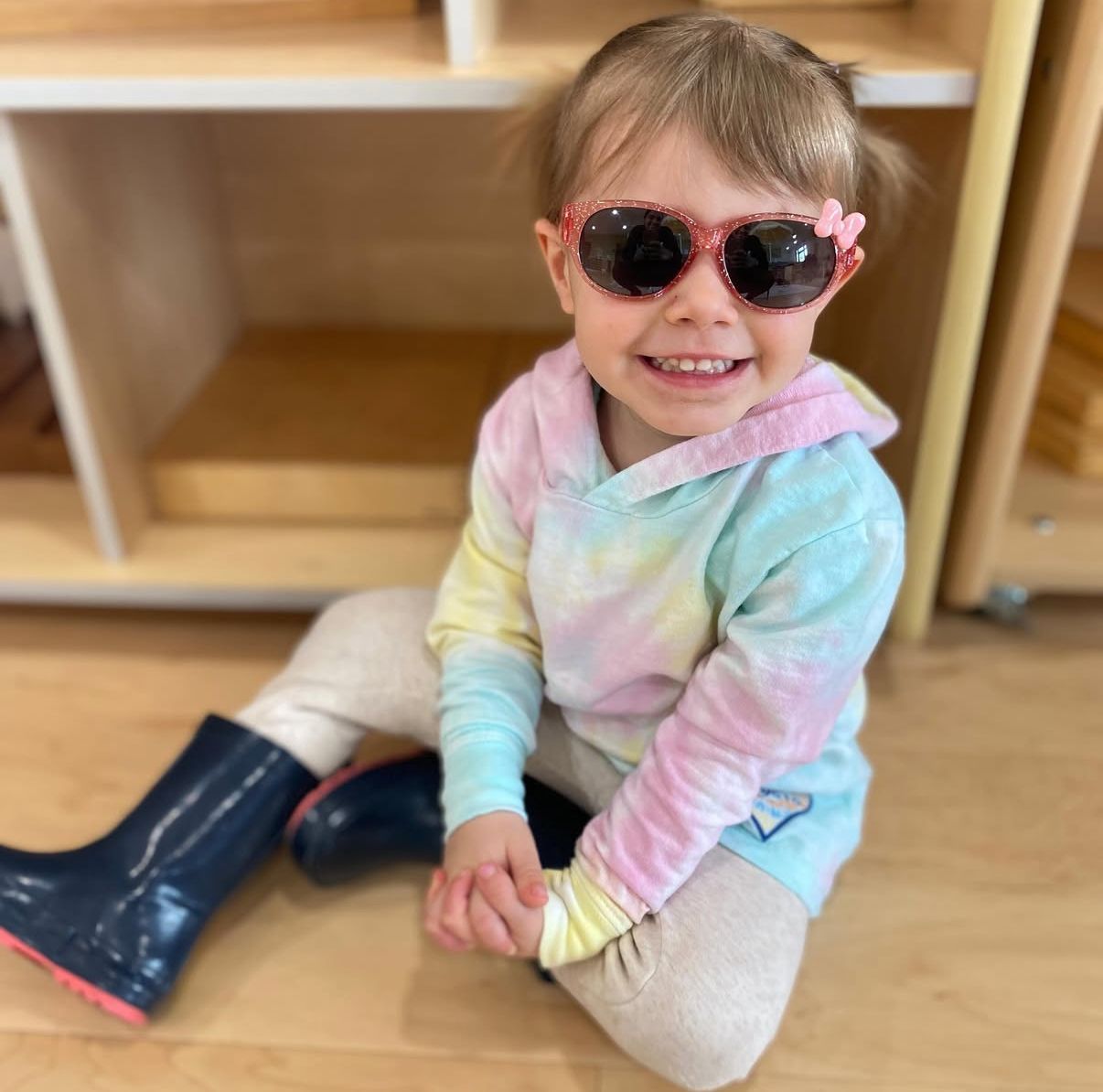 Girl wearing a Frozen helmet and pads, riding a purple scooter on pavement; another child scooters in background.