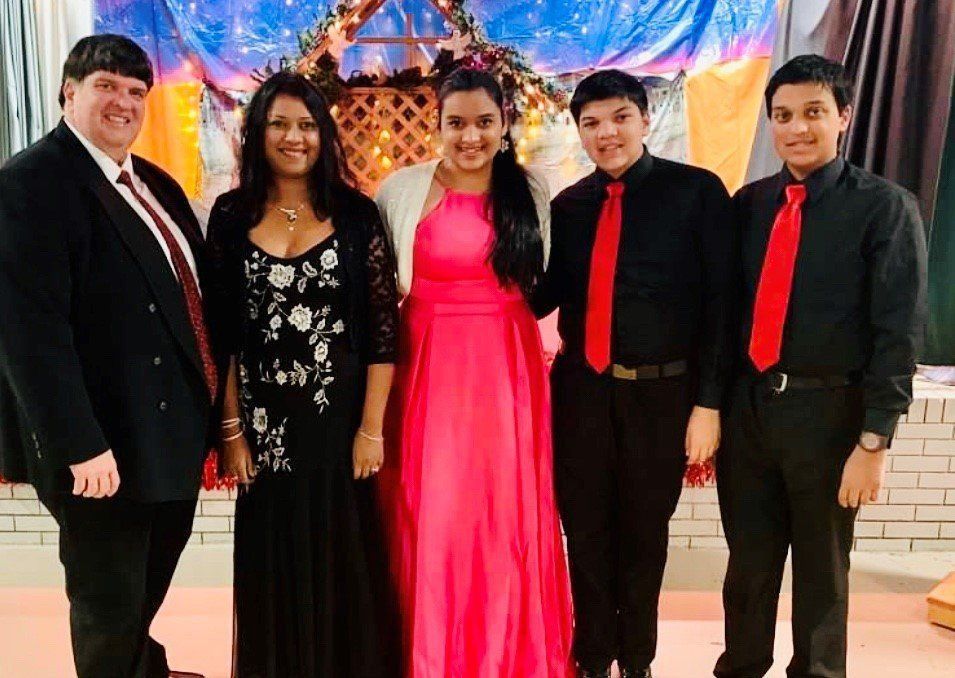 Five people posing formally: woman in pink dress, others in black and red, festive background.