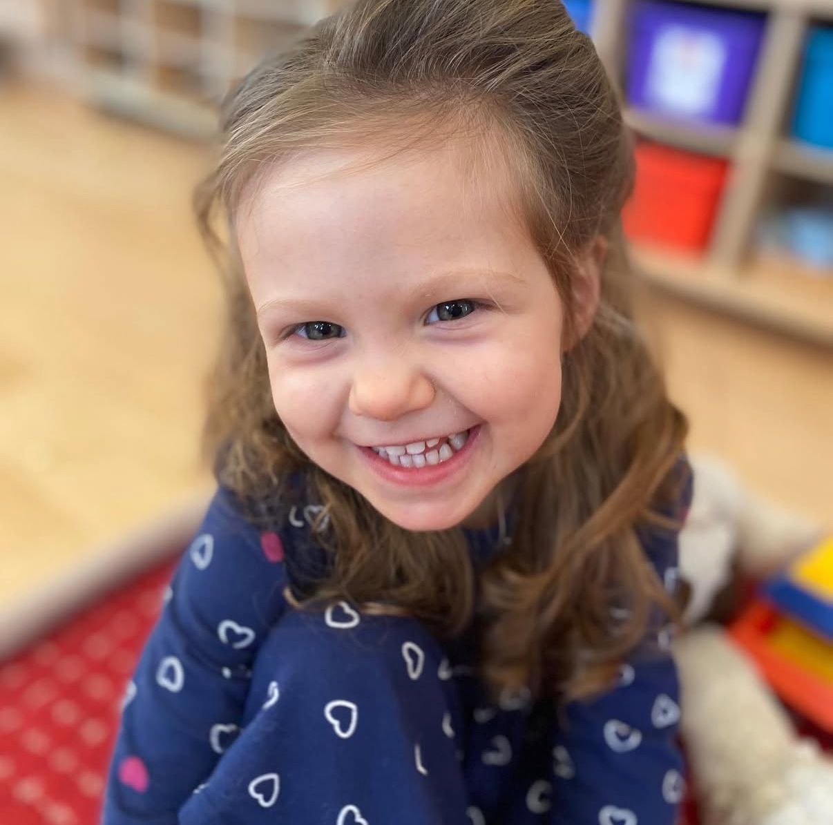 Girl in a blue top with heart patterns, smiling. Sitting on a red mat, room with colorful boxes in the background.