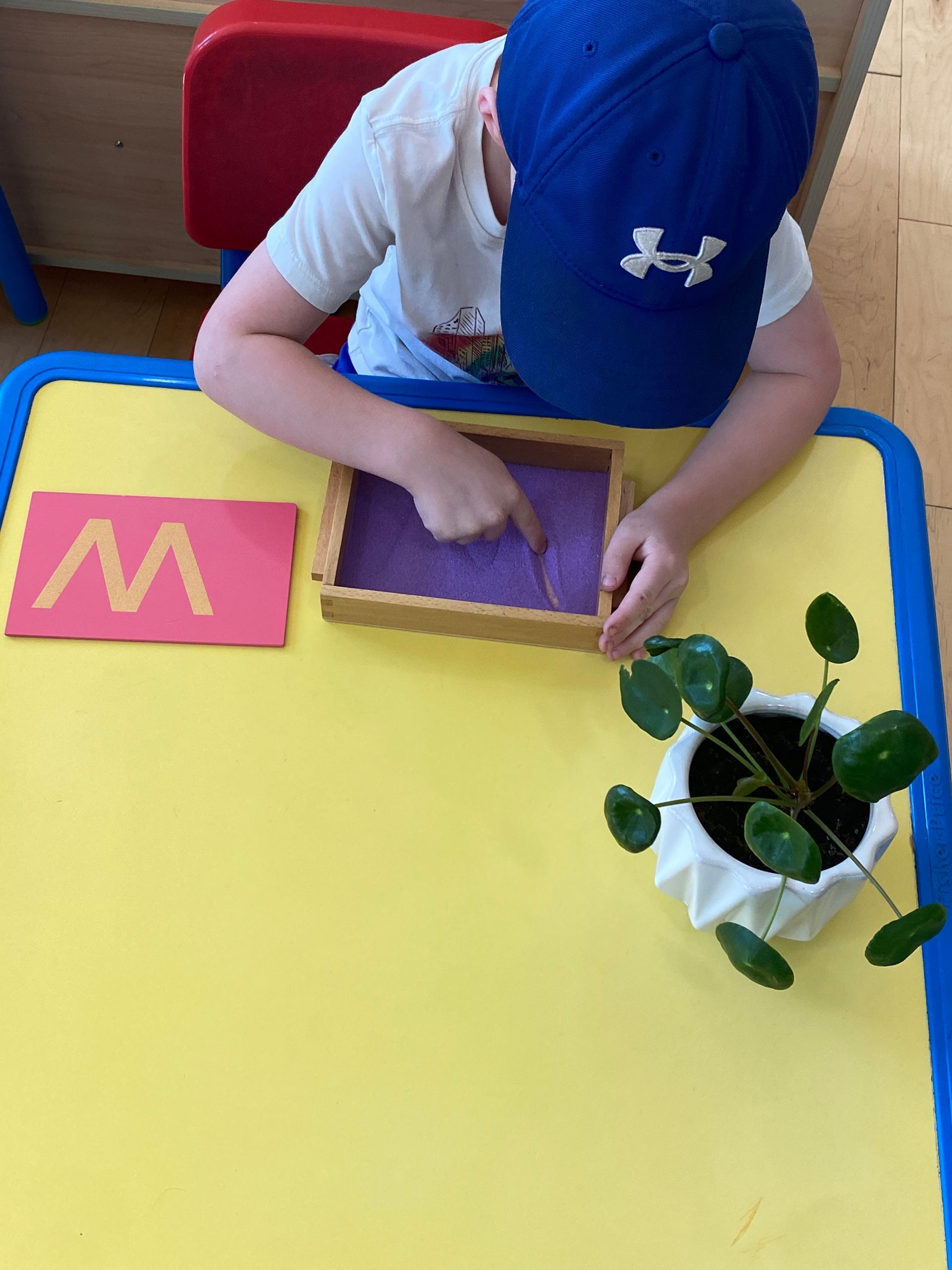 Child wearing blue cap traces lines in sand with finger, next to pink letter 