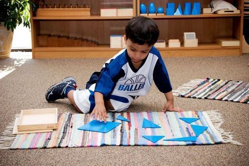 Boy on floor arranging blue geometric shapes on a rug in a classroom setting.