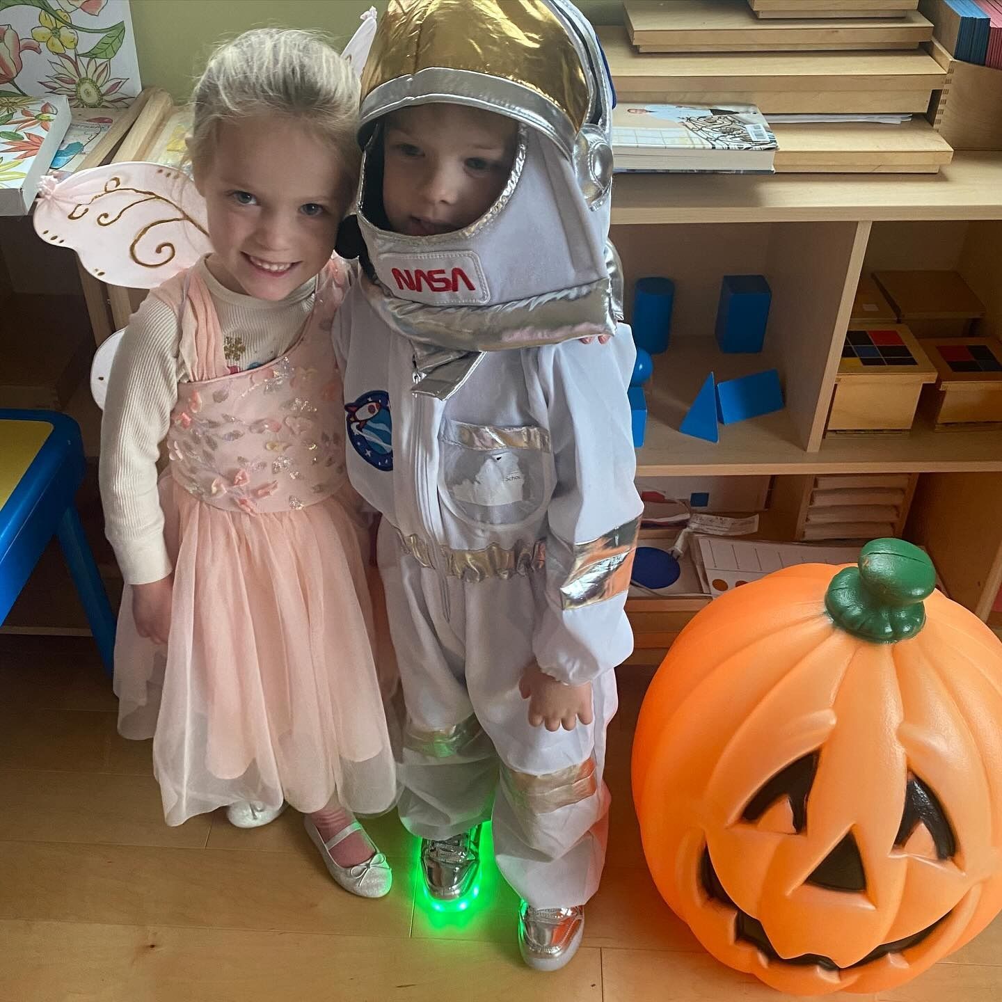 Girl in fairy costume and boy in astronaut suit beside a jack-o'-lantern, smiling.