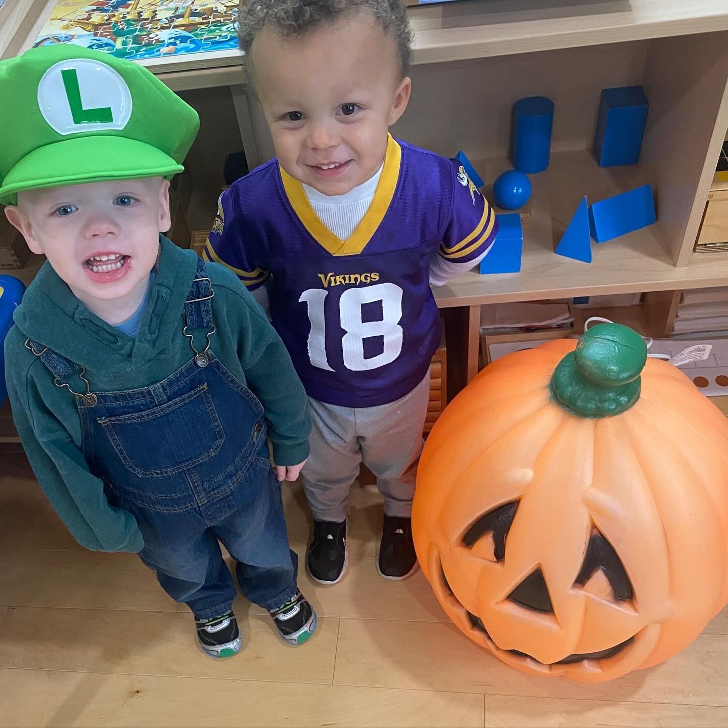 Two children dressed in costumes stand near a large plastic jack-o'-lantern. One wears a Luigi hat and overalls, the other a Vikings jersey.