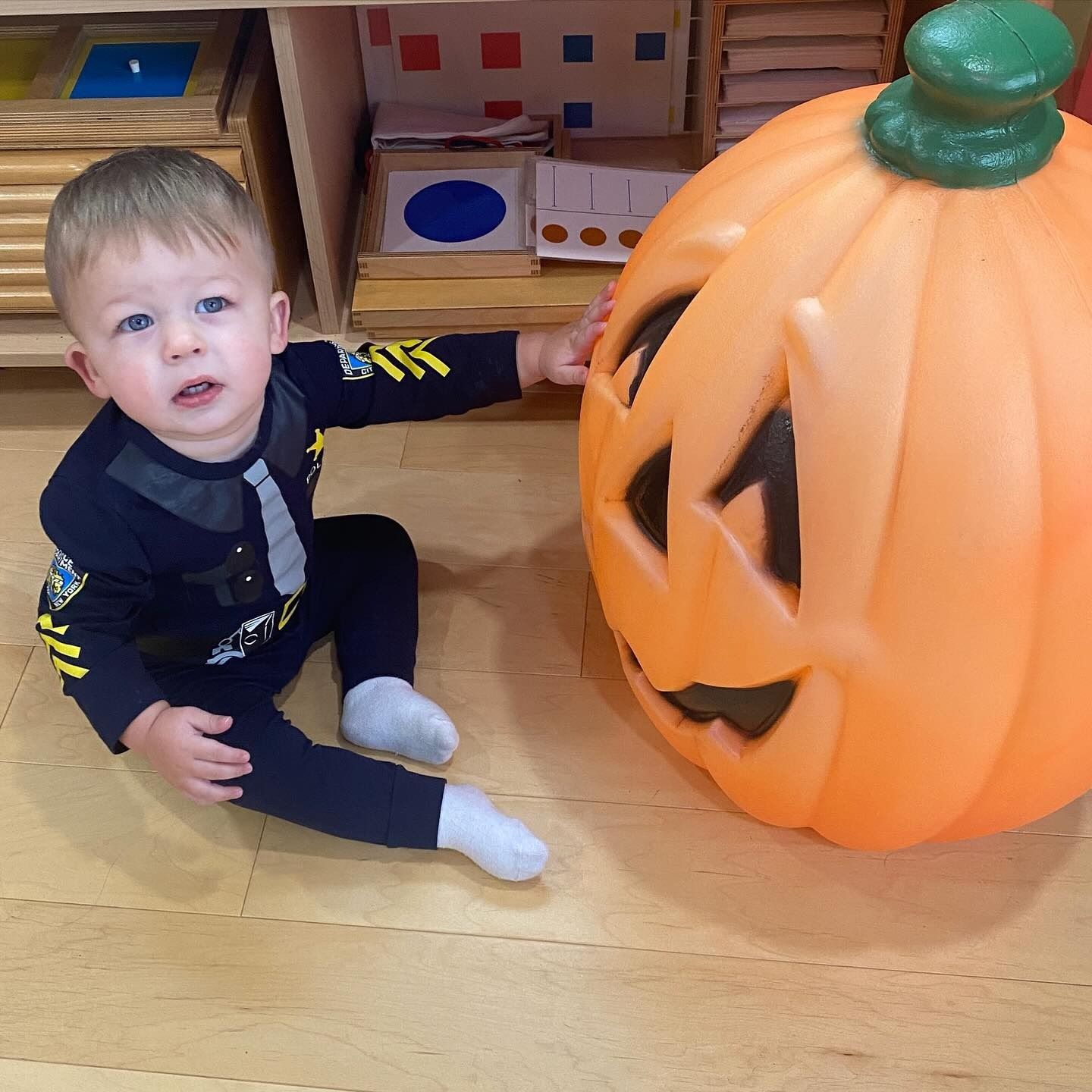Child in costume touches a large plastic jack-o'-lantern on wooden floor; child looks up.