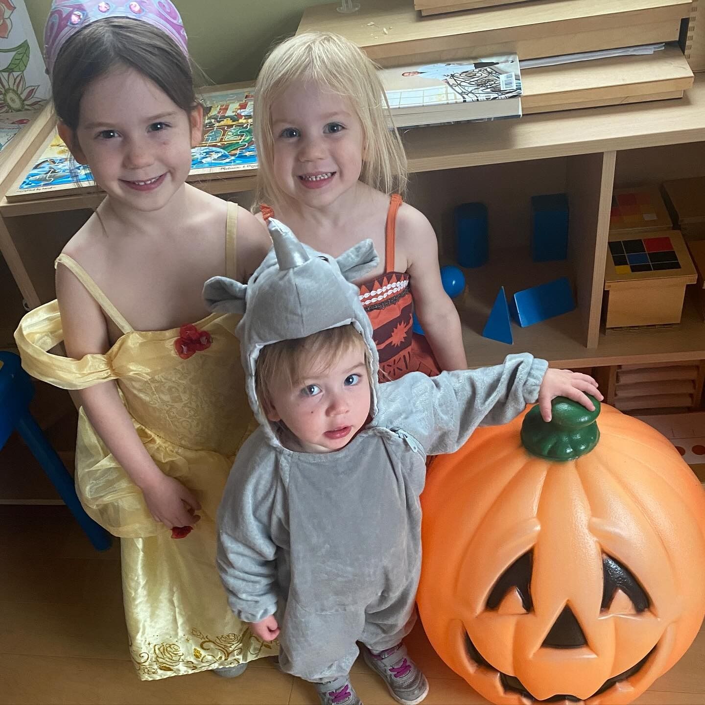 Three children in costumes smiling near a large pumpkin; a girl in a princess dress, one in a rhino costume.