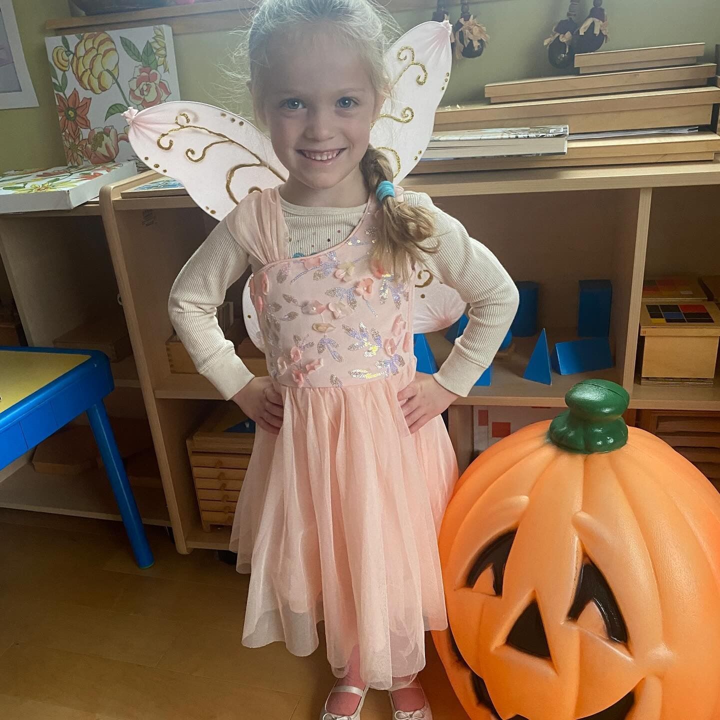 Girl in fairy costume smiles, posing with hands on hips, near a jack-o'-lantern and shelves.