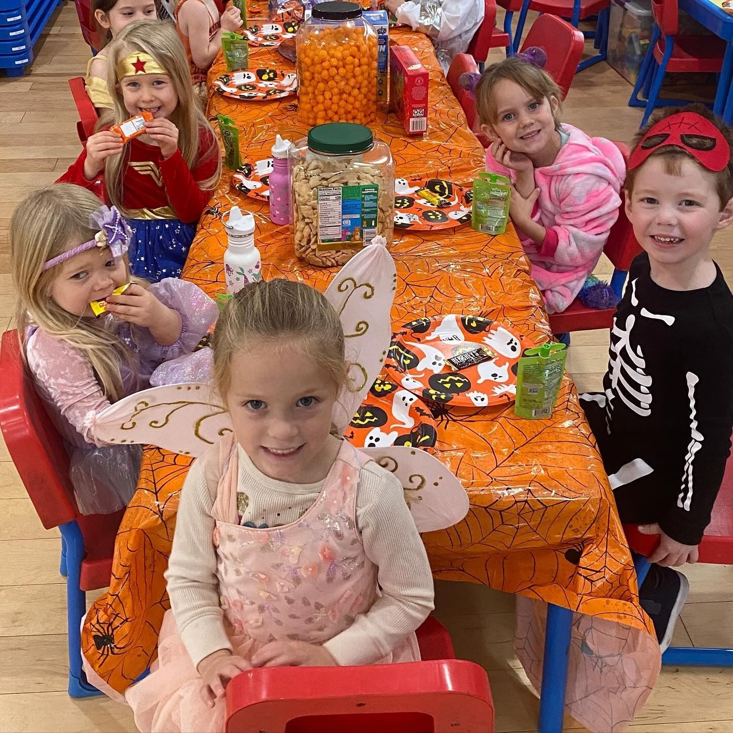 Children in costumes at a Halloween party, seated at a table with orange decorations and snacks.