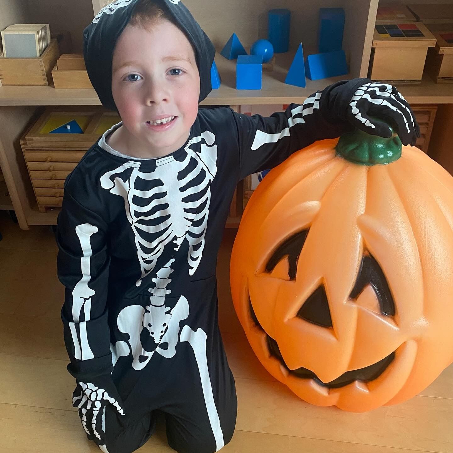 Boy in skeleton costume kneels next to a jack-o-lantern, smiling. Halloween setting.