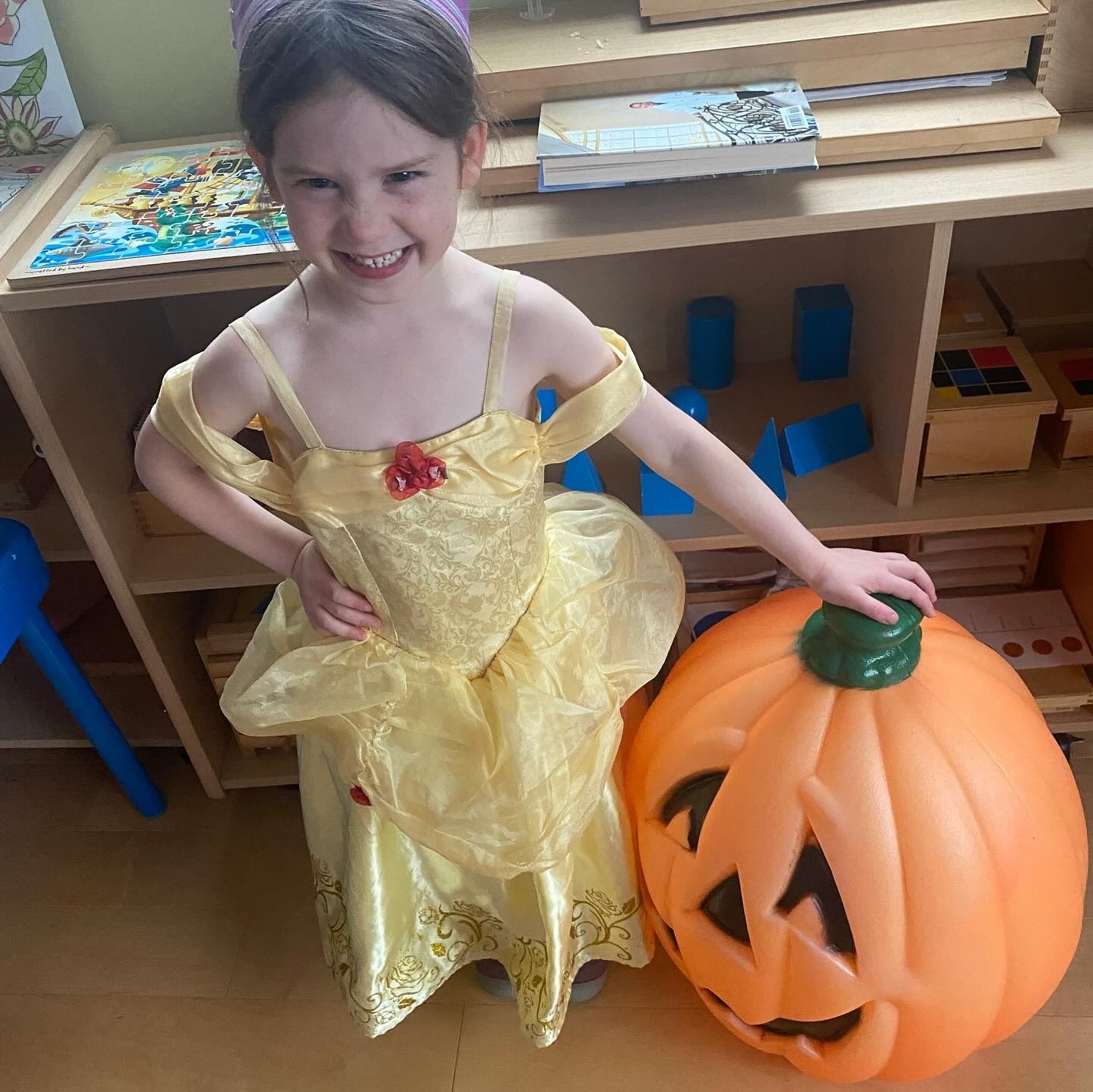 Girl in yellow dress, holding pumpkin. She is in a room with shelves. She is smiling.