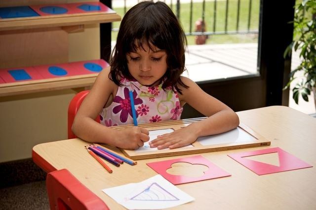 Child drawing at a desk with colored pencils, shape stencils, and a shape sorting activity in the background.