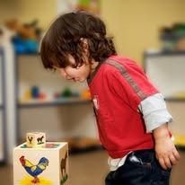 Child looking at a toy block, wearing a red shirt, in a playroom setting.