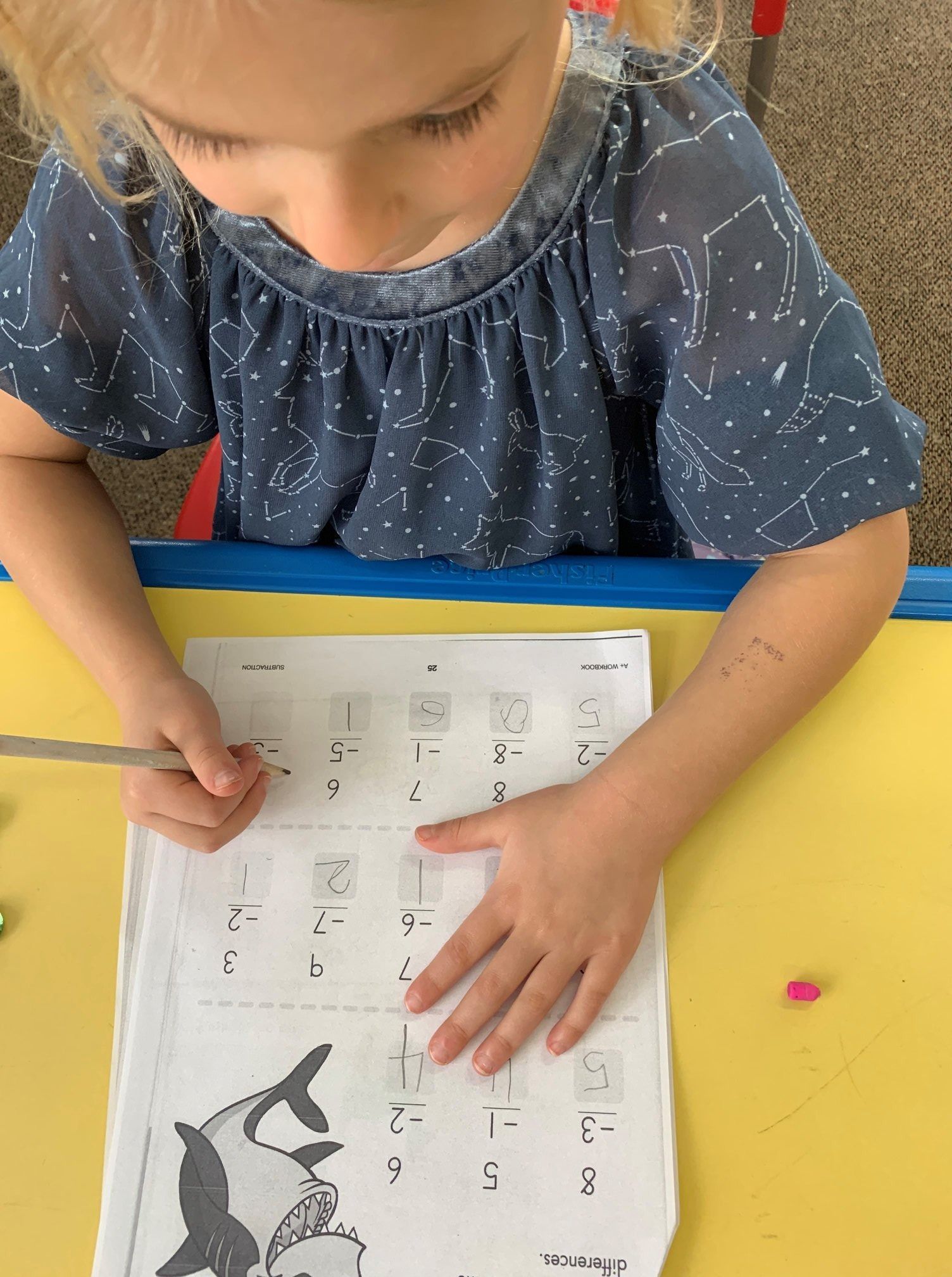 Child in blue shirt writing on worksheet with a drawing of a shark, at a yellow desk.