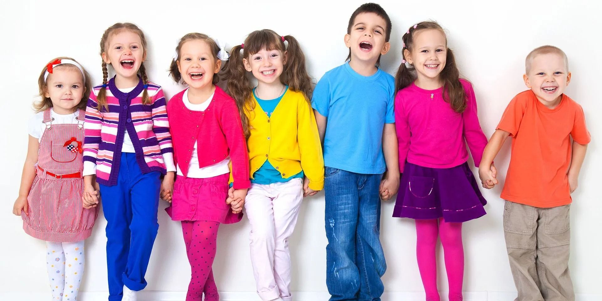 Seven children smiling, holding hands, in front of a white wall. They wear colorful clothes.
