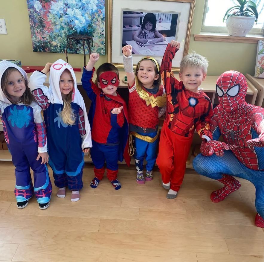 Children in superhero costumes pose with an adult in a Spider-Man suit indoors.