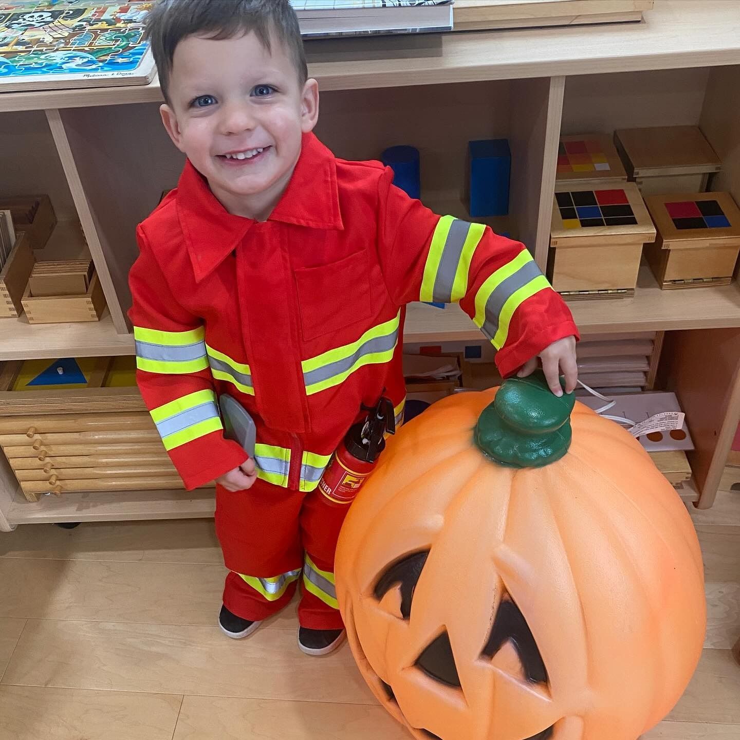 Boy in firefighter costume smiles next to a jack-o-lantern in a classroom.