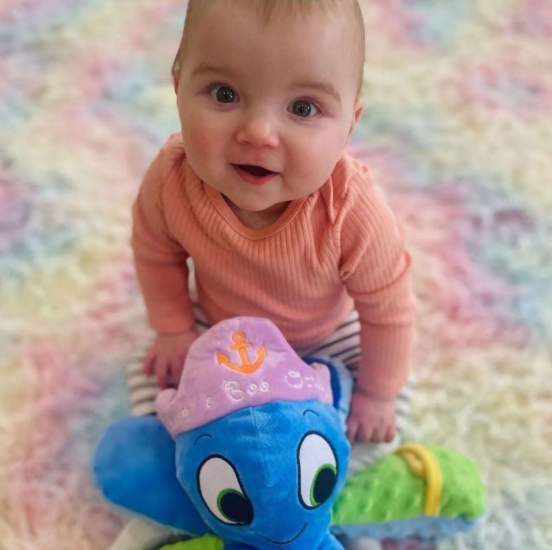 Baby smiling, seated with a blue octopus toy wearing a pink pirate hat on a multi-colored rug.