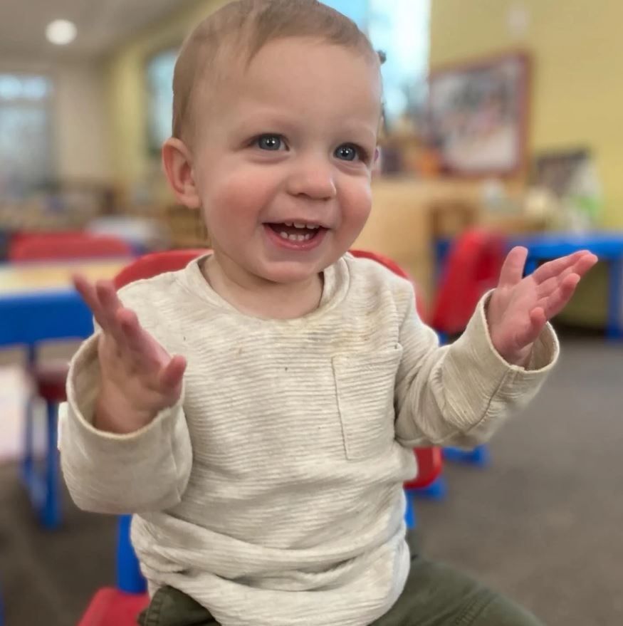 Happy toddler clapping with hands raised, sitting in a red chair. Beige shirt, green pants, classroom background.