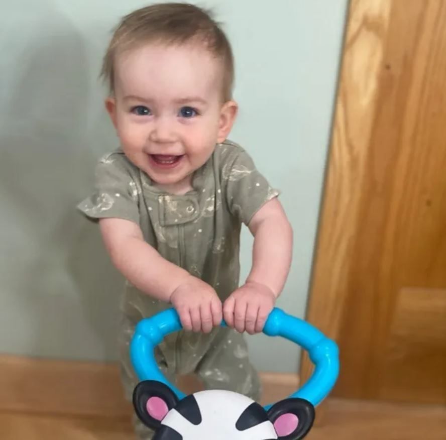 Smiling baby in green jumpsuit, holding a blue and black walker in front of a blue and wooden wall.