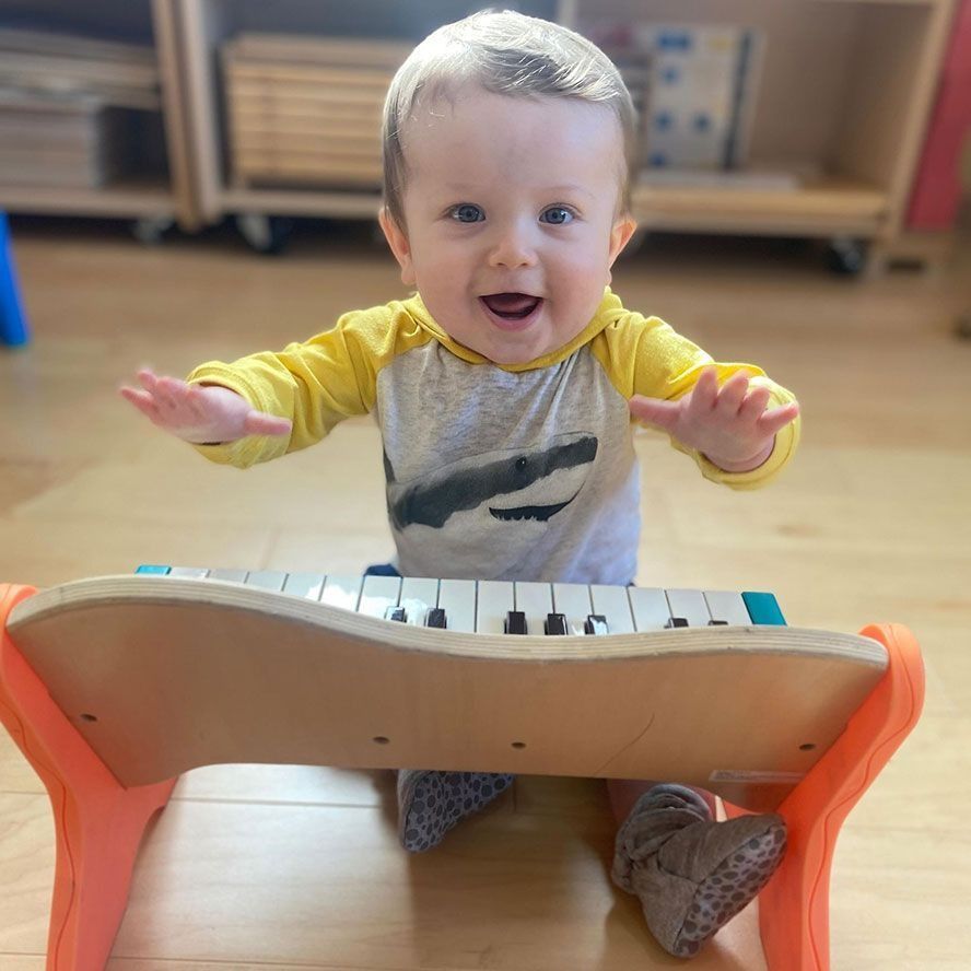 Smiling baby with arms raised, playing a toy piano. Indoors, wooden floor, wearing a yellow hooded shirt.