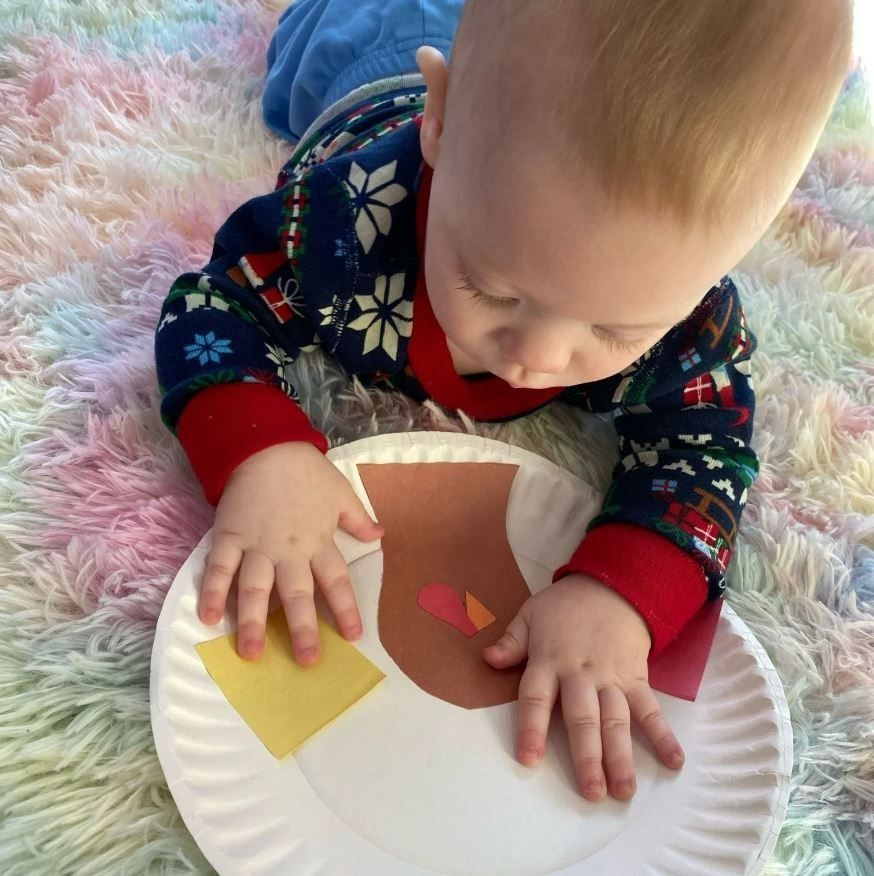 Baby on fuzzy rug playing with paper plate decorated with colorful paper pieces.