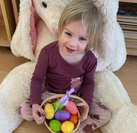 Girl holding a basket of Easter eggs, smiling in front of a giant bunny, wearing a burgundy shirt and patterned pants.