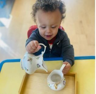 Toddler pours from a floral teapot into a matching creamer on a tray.