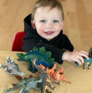 Young child with rosy cheeks smiles while playing with toy dinosaurs on a wooden table.