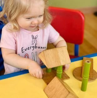 Child plays with wooden blocks at a yellow table; red chair in background.