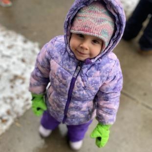 Child wearing purple winter coat and hat, green gloves, and purple pants smiles outside.