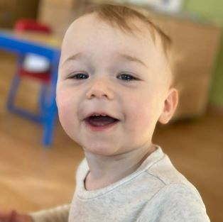 Smiling toddler indoors, looking at camera. Blue table and chair in background.