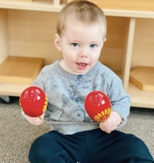 Toddler holding red maracas, smiling, sitting on a rug indoors.