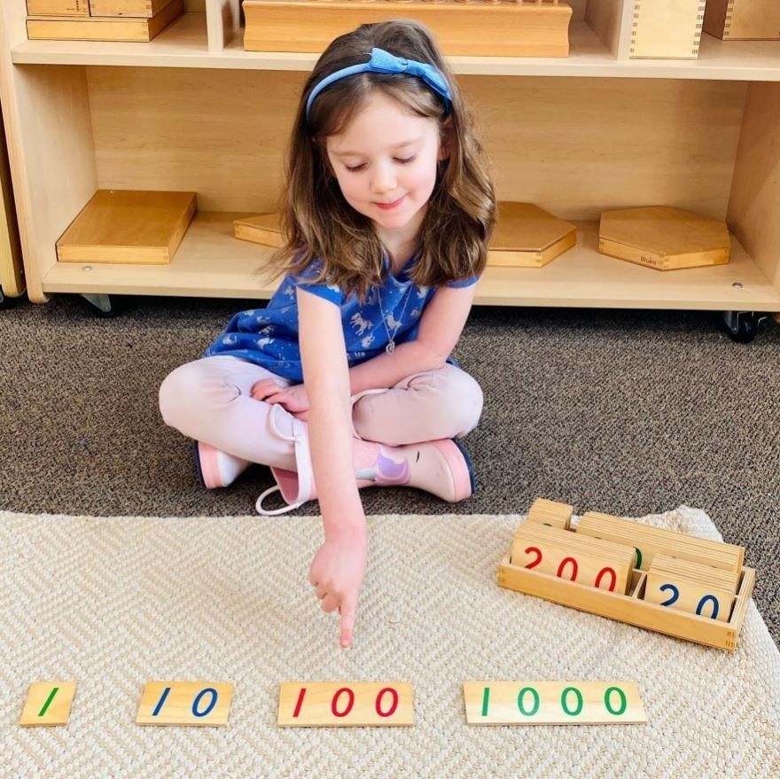 Young child pointing at Montessori math materials: 1, 10, 100, 1000. Child is smiling.