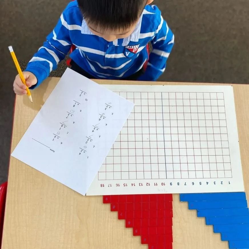 Young child in blue stripes writing on paper next to a grid and colored blocks on a table.