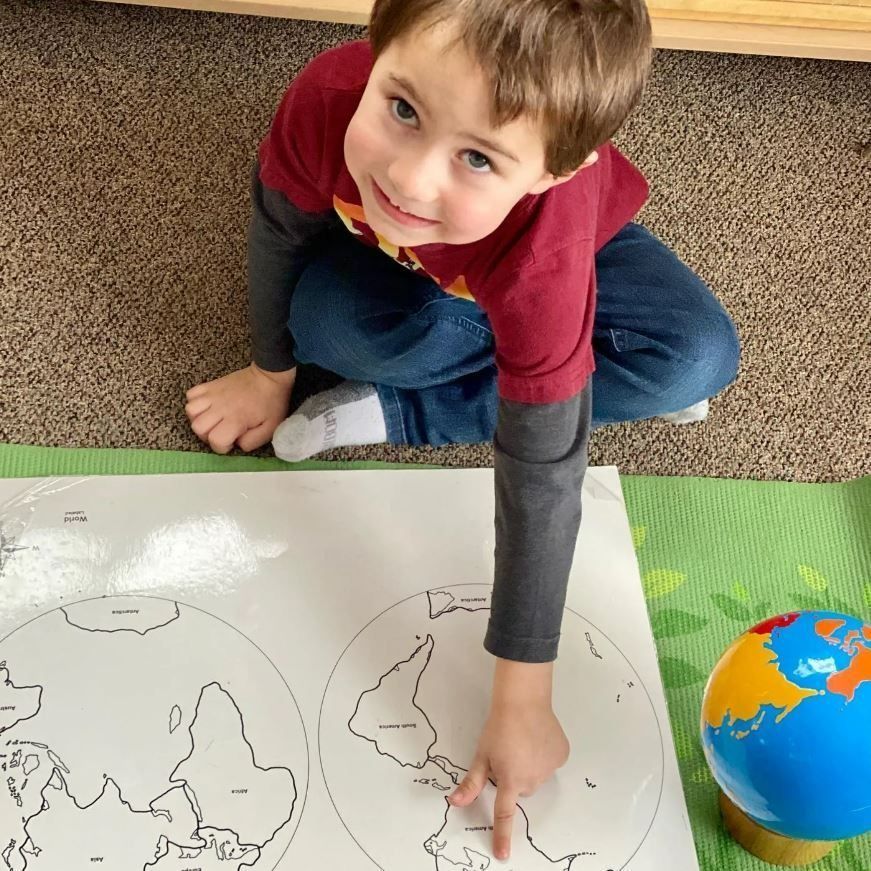 Young child pointing at map on floor next to globe. Child smiles, wears red/gray shirt and jeans.